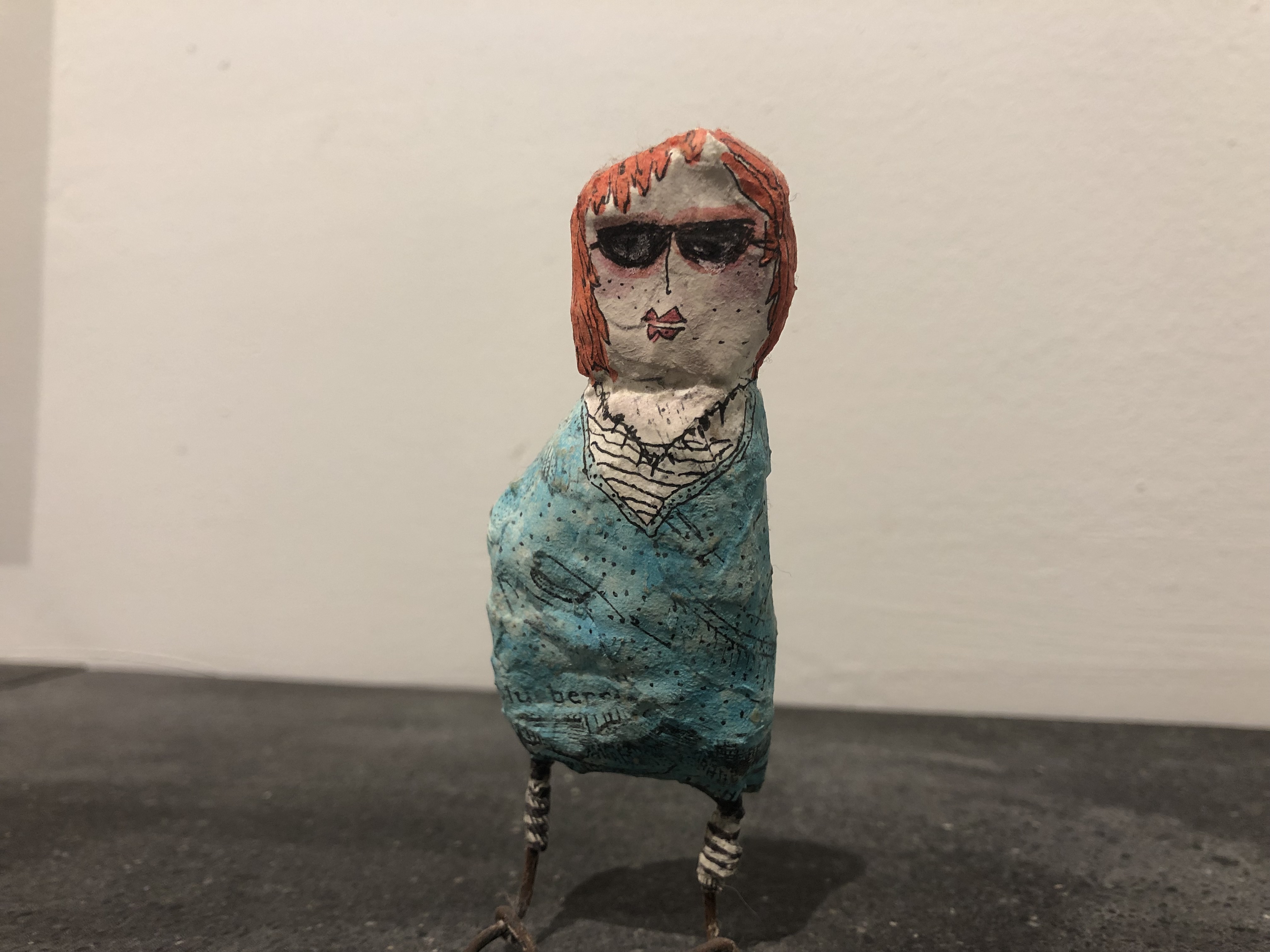 Paper-mache person with orange hair, sunglasses, and teal sweater.
