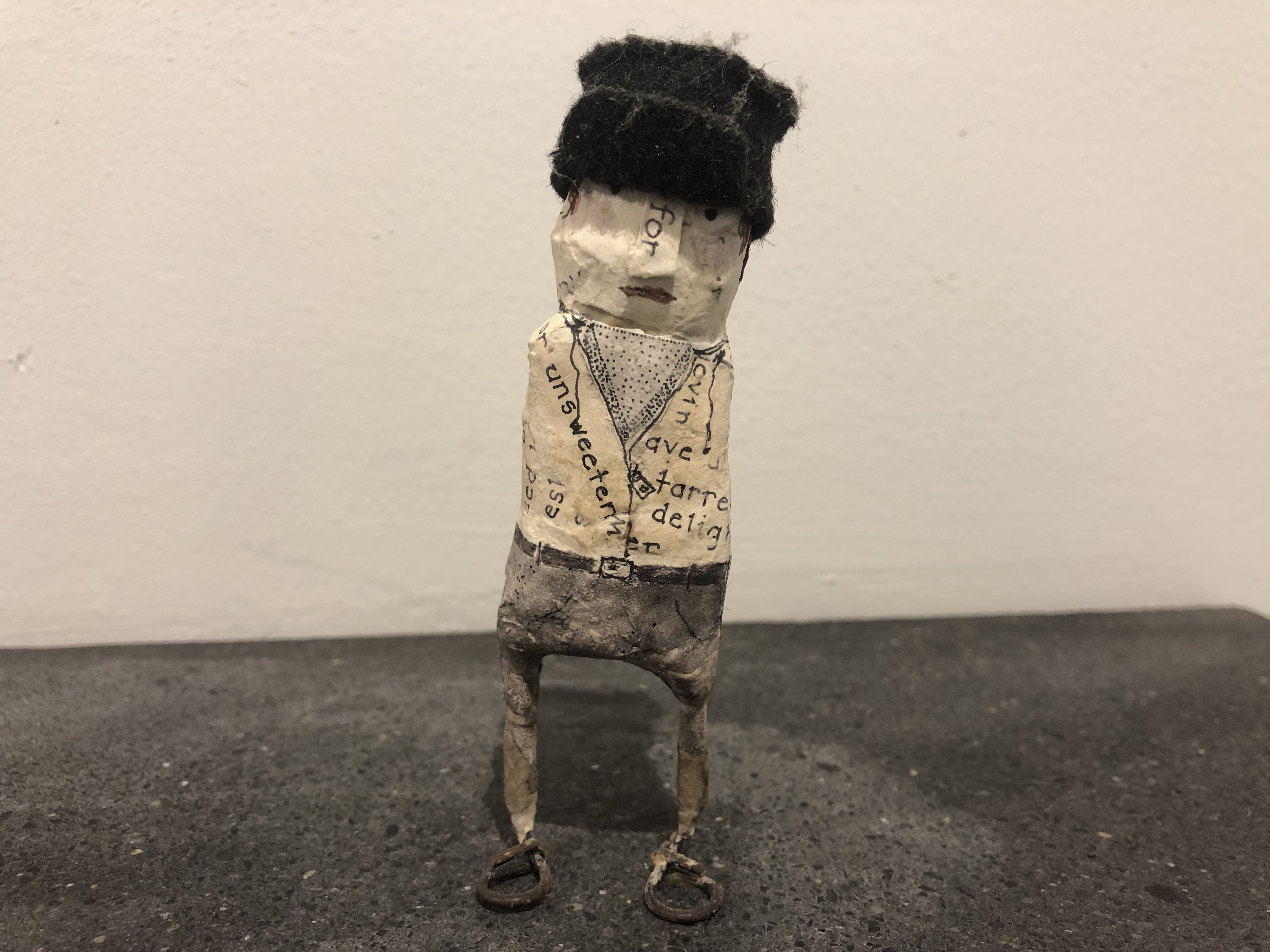 Paper-mache person with fuzzy hat, tucked in shirt, and grey pants.